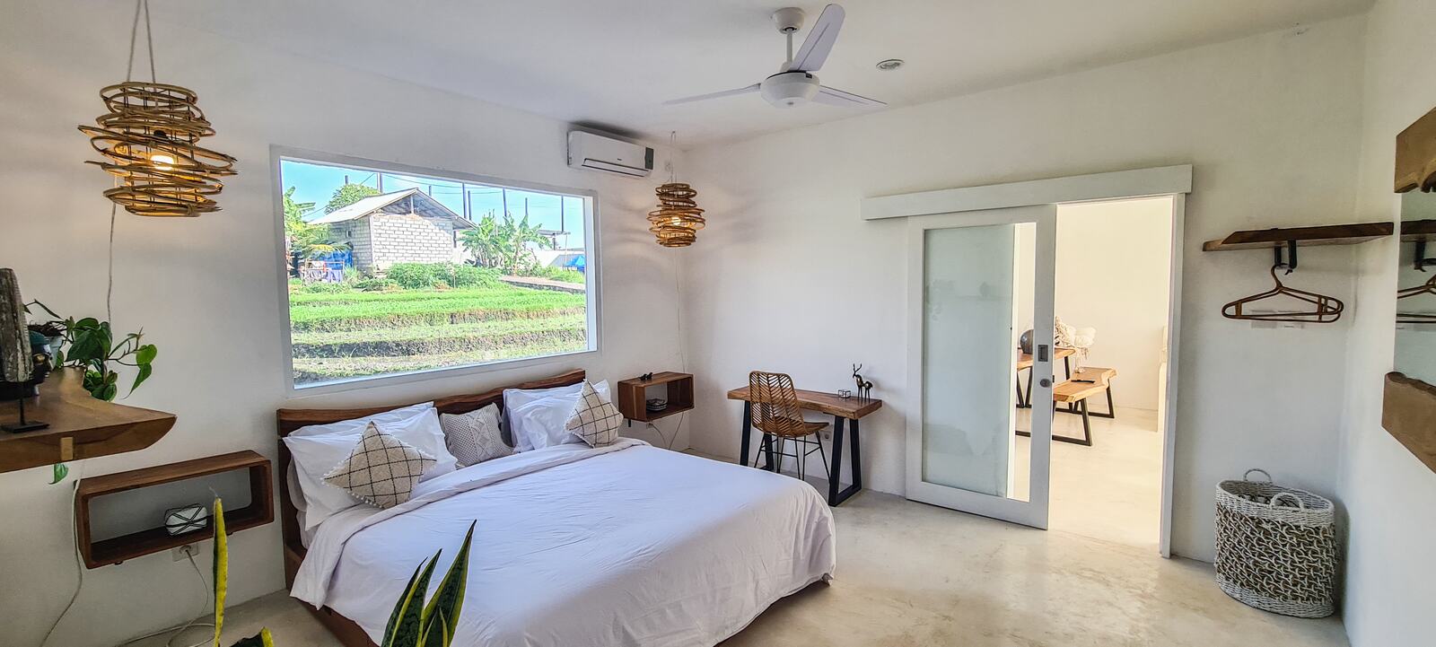 Bedroom with a king bed in white linen, a wide picture window, woven pendant lamps and a teak work desk