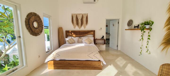Queen bedroom with a teak bed, round rattan mirror, macrame hanging and a tall window onto frangipani leaves