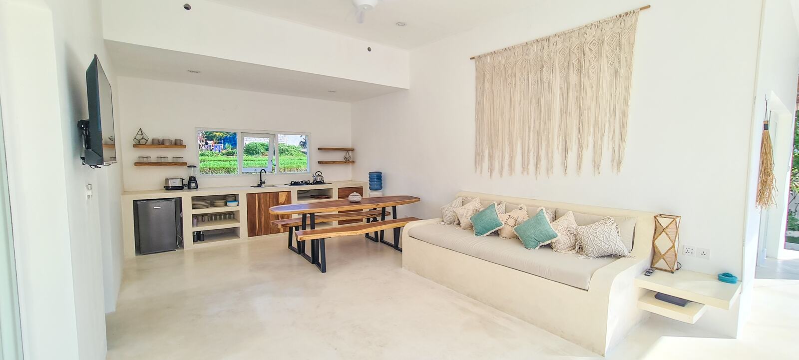 Open-plan living room with a long white built-in sofa, macrame wall hanging, teak dining table and the kitchen behind